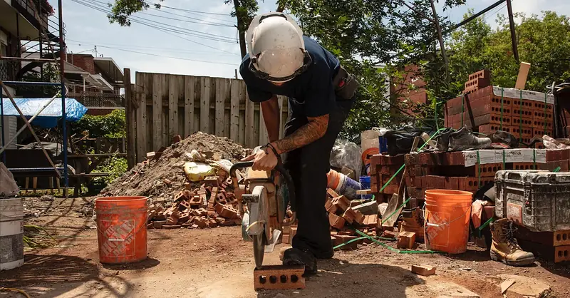 Maçon de Maçonnerie JP inc., casque et visière, qui coupe une brique à la scie sur le chantier
